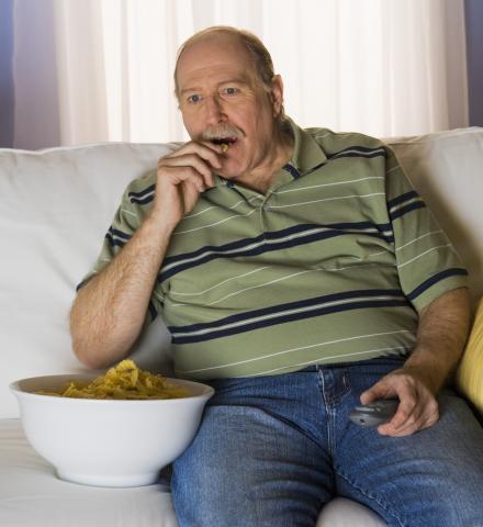 Imagen de un hombre viendo la televisión y comiendo patatas fritas, como ejemplo de comportamiento sedentario.