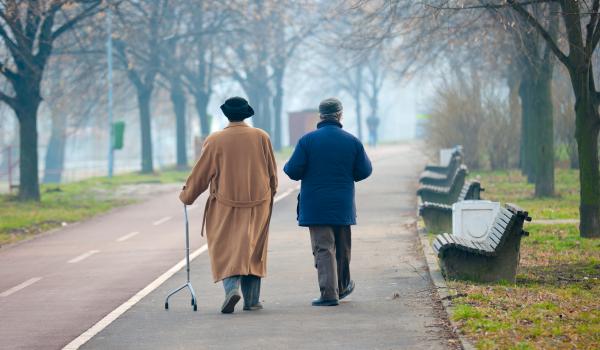 Two older people walking outdoors. One of the two uses a walking aid, i.e. a three point cane