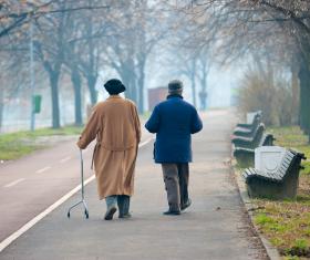 Two older people walking outdoors. One of the two uses a walking aid, i.e. a three point cane